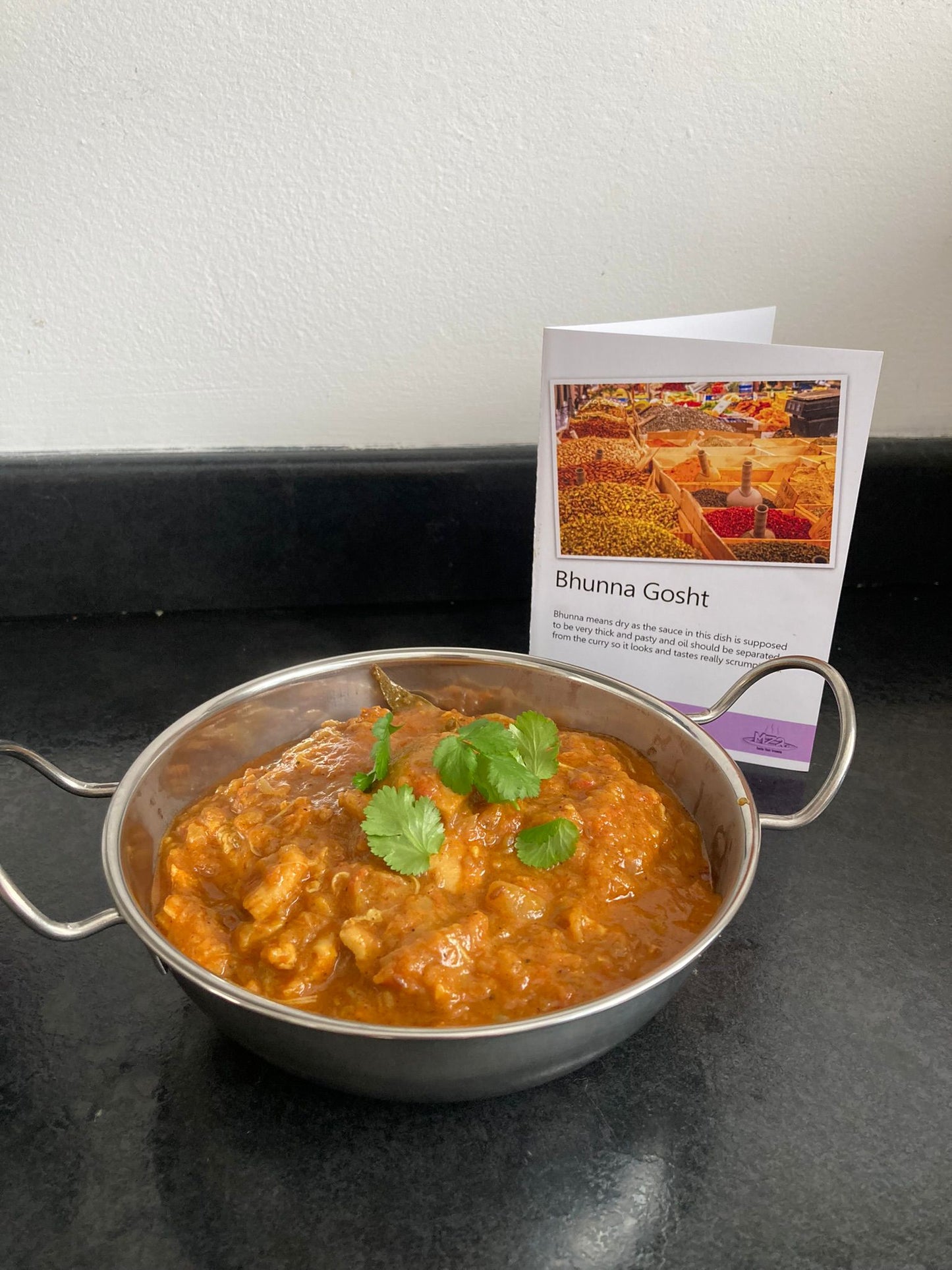 Bhunna Gosht in a balti bowl with the recipe card behind it, all on a table.