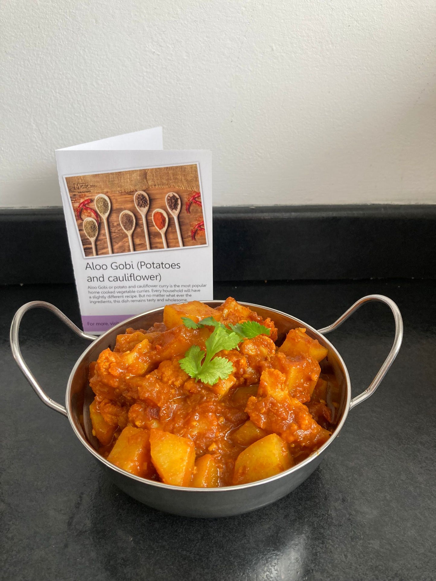 Aloo Gobi in a balti dish with the recipe card behind it.