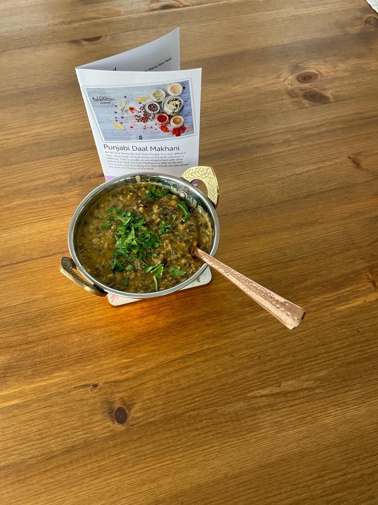 a slightly distant shot of punjabi daal makhani in a metallic copper coloured bowl with a spoon sticking out of it. Behind it is its recipe card, all placed on a wooden table.