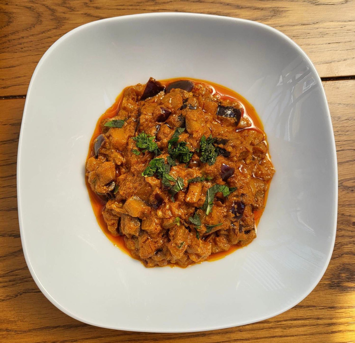 Photo of Bengan Bharta Aubergine Curry centred artistically in the middle of a white, rounded square shallow bowl.